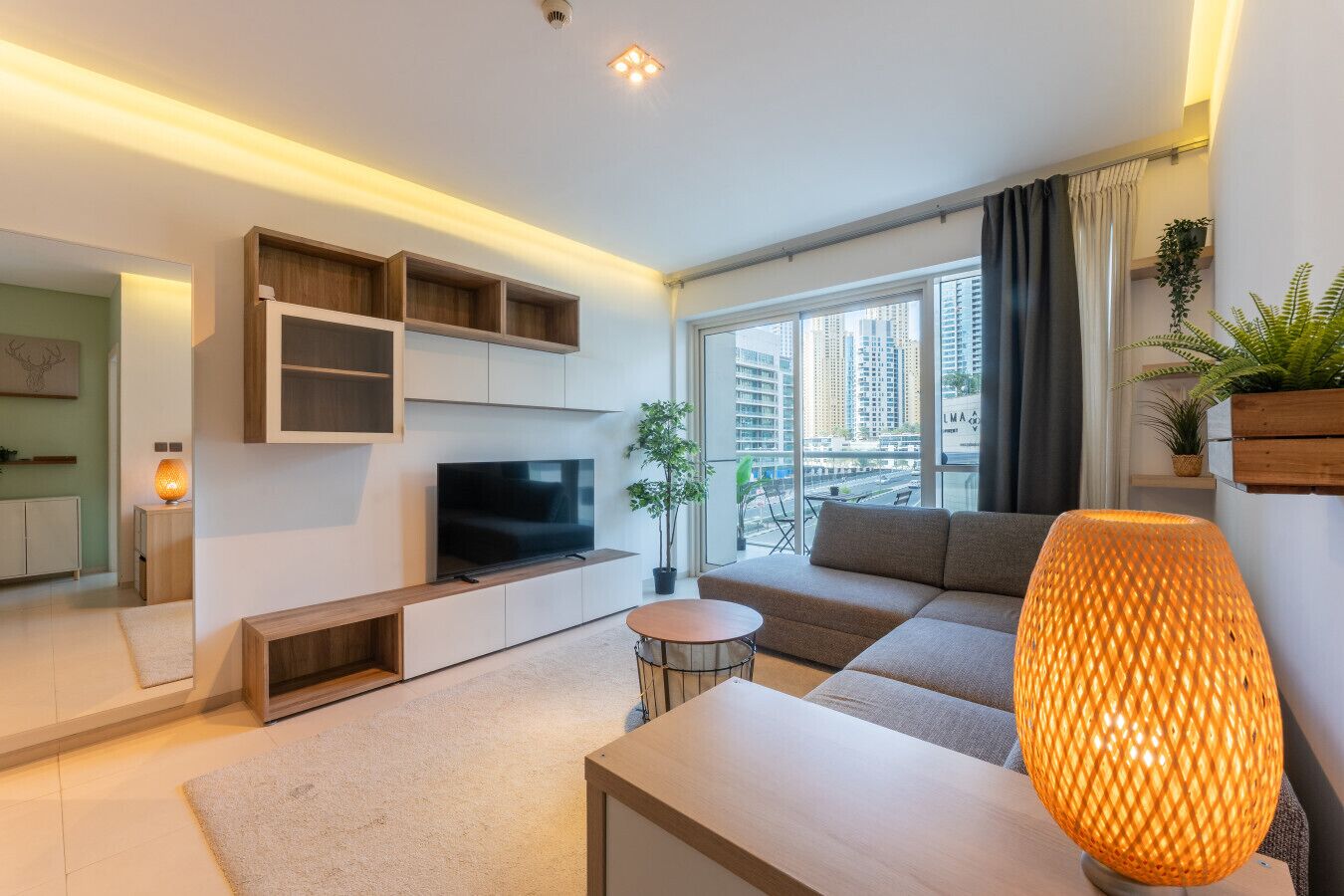 A modern living room with a flat-screen TV, a sofa, a coffee table, and a large window offering a city view.