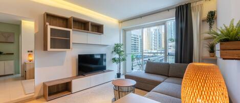 A modern living room with a flat-screen TV, a sofa, a coffee table, and a large window offering a city view.