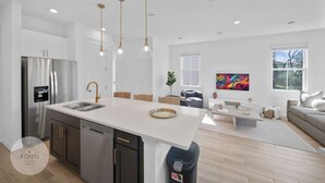 A modern kitchen with a central island, stainless steel appliances, pendant lighting, and a view to a living area with a sofa and a colorful painting. - 1052 3BR Luxury Resort House by Disney Orlando (Davenport)