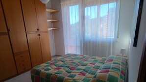 Ein Schlafzimmer mit einem Bett, einem Holzschrank, einem kleinen Tisch mit Lampe und einem Fenster mit transparenten Vorhängen.