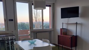 Ein Essbereich mit Tisch und Stühlen, einem Fernseher auf einem Schrank und Blick durch große Fenster.