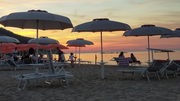 Ein Strand mit mehreren Sonnenschirmen und Liegestühlen, der auf einen Strandhotelkomplex hindeutet.