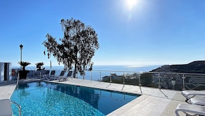 Cielo azul despejado, piscina con vistas al mar y sillones de descanso.