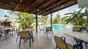 A covered outdoor seating area with tables and chairs, a pool, and tropical plants.