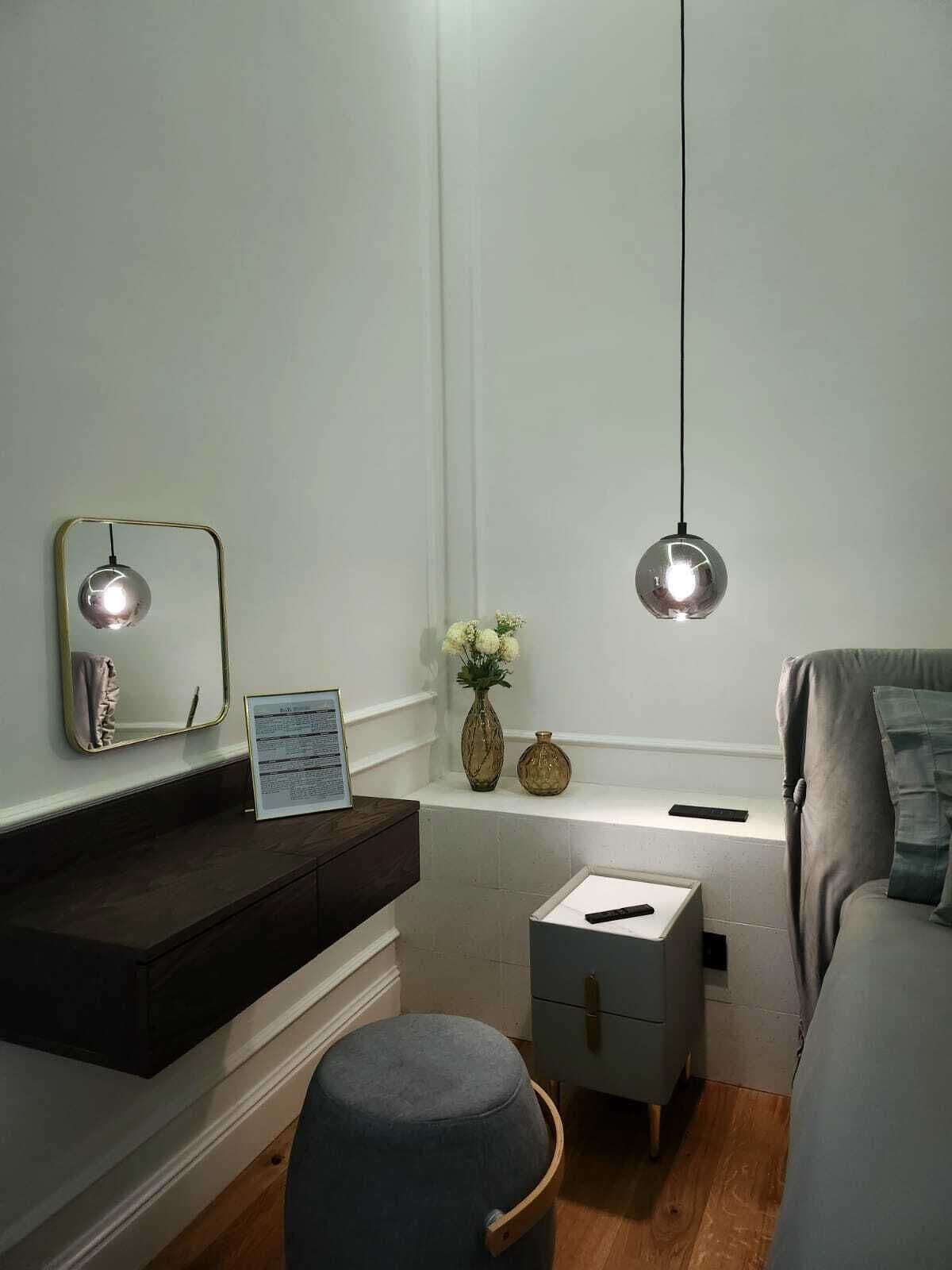A modern bedroom with a dark wooden dresser, a small bedside table, a grey ottoman, and a grey sofa.