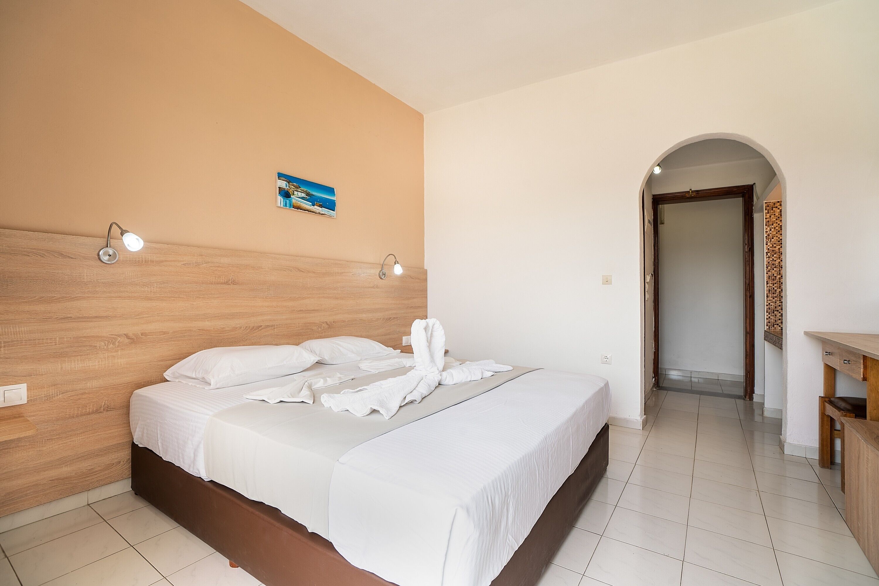 A hotel room with a wooden headboard, a bed with white linens, two bedside lamps, and a small painting on the wall.