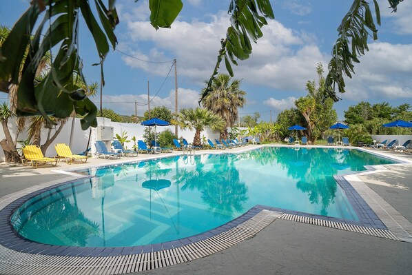 Une piscine entourée de chaises longues et de parasols, bordée de palmiers et surplombée d’un ciel dégagé.