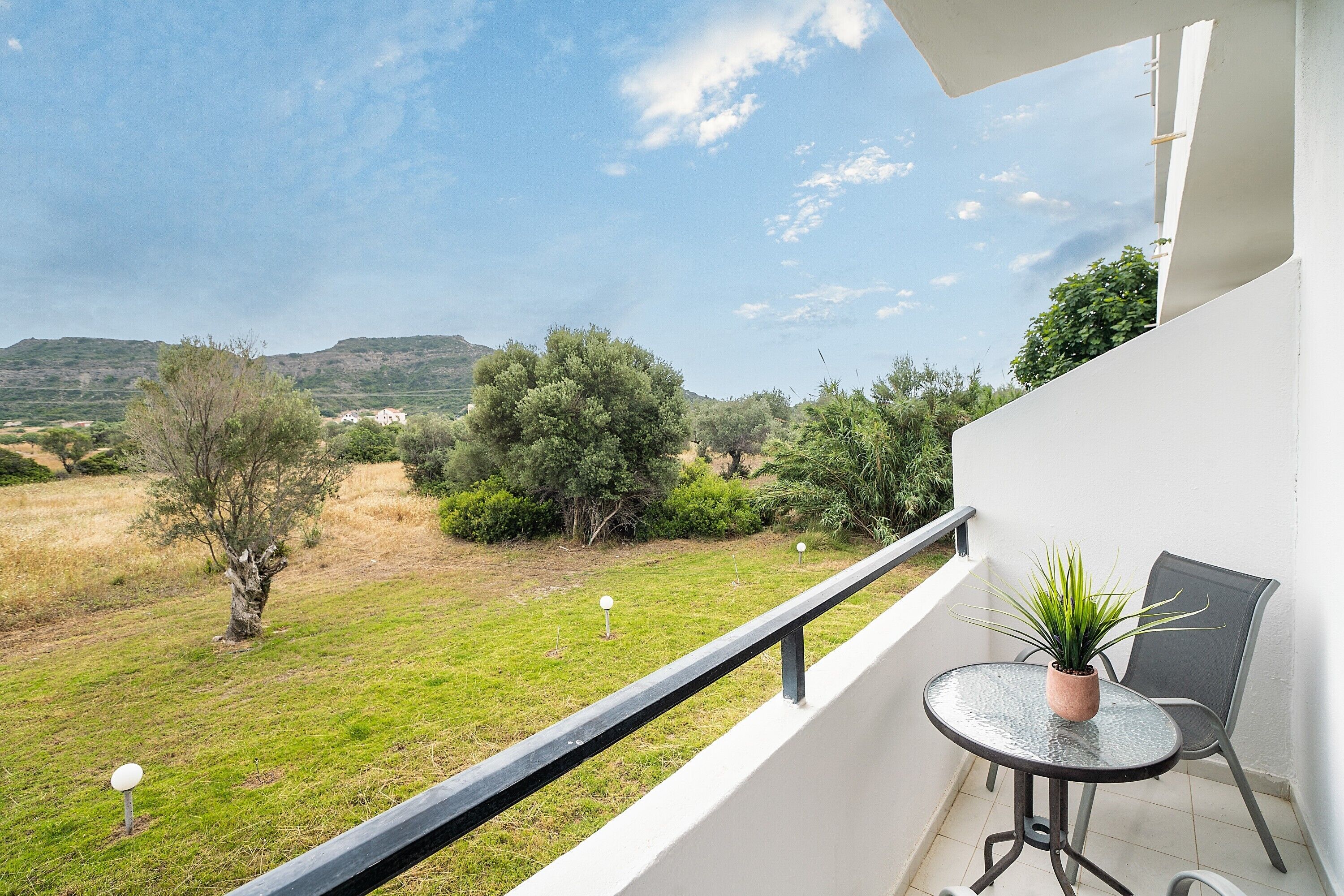 A balcony with a potted plant, a chair, and a view of a grassy landscape with trees and hills.