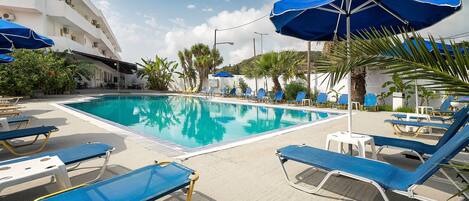 A pool area with blue sun loungers and umbrellas, a clear pool, and a white building with balconies.
