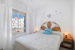 A bedroom with a bed, a nightstand with a lamp, a window with a view of buildings, and a wall decal with stars and a moon.