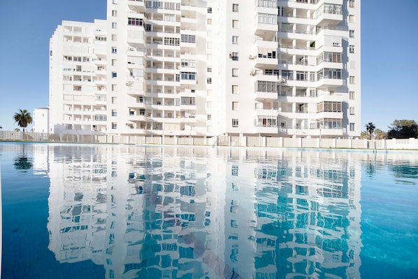 A high-rise building with multiple balconies and a clear blue pool in front.