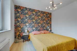 Une chambre à coucher avec un papier peint à motifs floraux, un lit avec une literie jaune, une table de chevet avec une lampe et un lustre moderne.