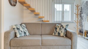 A living room with a sofa, patterned cushions, a wooden staircase, and a window. - Holiday Home 'Le Banc Des Hermelles' with Private Terrace, Private Garden and Wi-Fi (Saint-Benoît-des-Ondes)