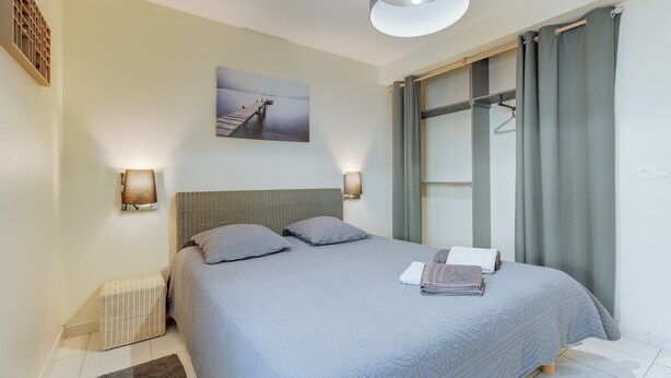 Un dormitorio con cama, cabecera, cuadro en la pared, lámpara de noche, perchero y una ventana con cortinas.