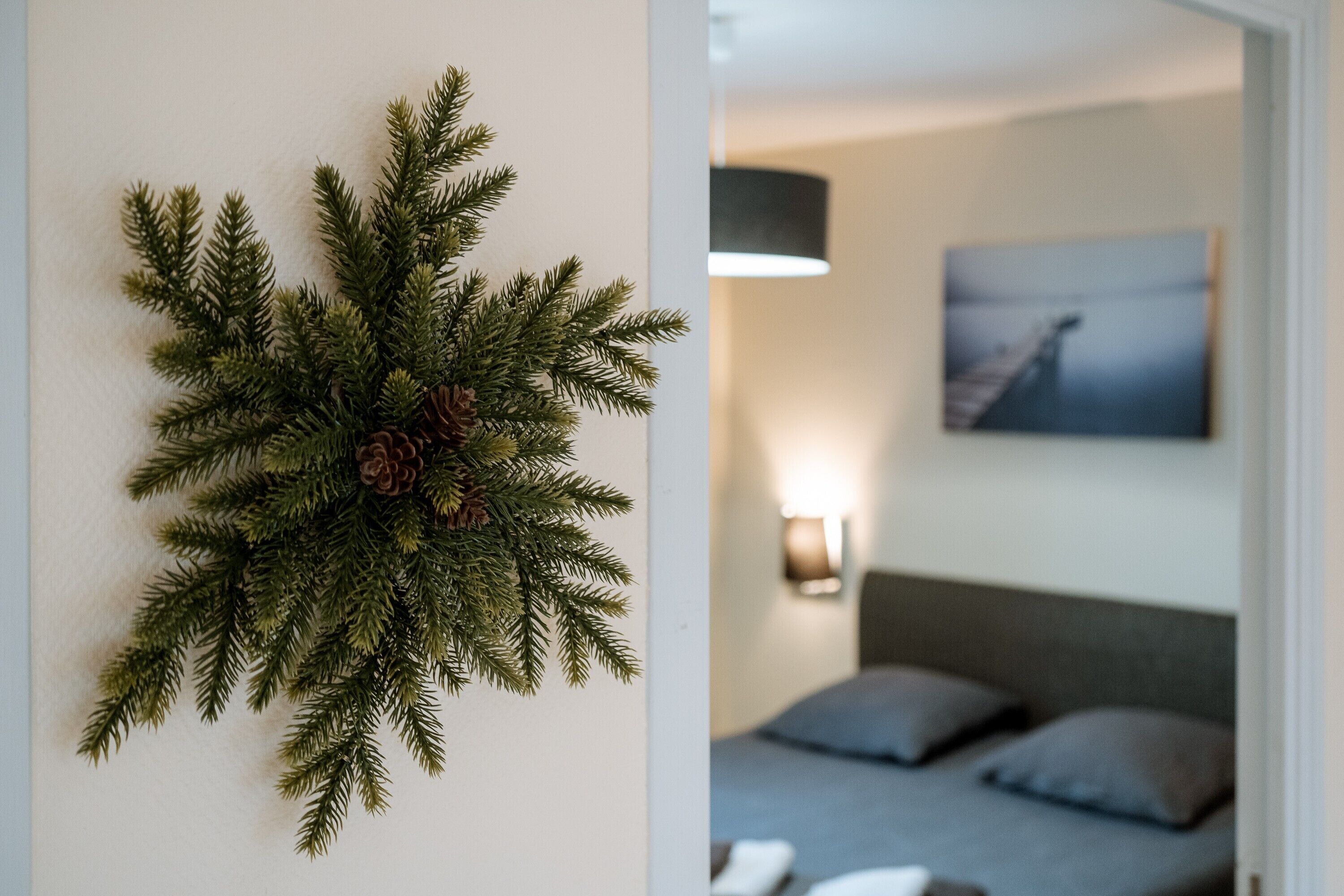Eine Wanddekoration aus Kiefernzweigen mit Zapfen, ein Bett mit grauer Bettwäsche und ein Gemälde an der Wand.