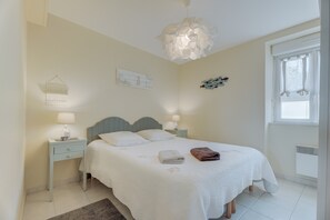 A neatly made bed with pillows, a bedside table with a lamp, a wall-mounted artwork, and a window with curtains.
