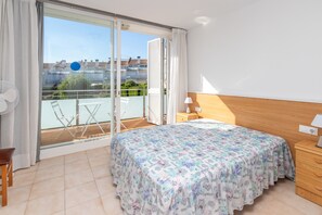 A bedroom with a bed, a wooden nightstand, a chair, a fan, a balcony with a table and chairs, and a view of buildings.
