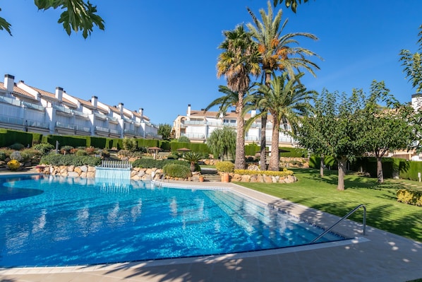 A swimming pool with a clear blue water, surrounded by a well-maintained lawn, palm trees, and a row of white buildings with balconies.