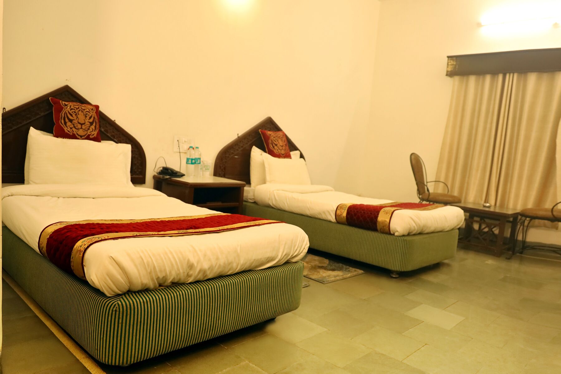 Two single beds with red and gold accents, a nightstand with a phone and water bottles, a chair, and a window with curtains.