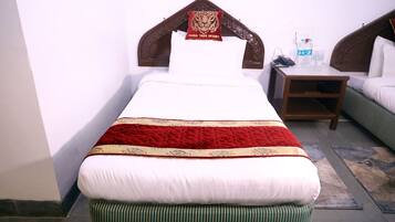 Uma cama de solteiro com um edredom vermelho e bege, uma cabeceira de madeira e um travesseiro com o logotipo de uma tigresa. Há uma pequena mesa de cabeceira de madeira com um telefone e garrafas de água ao lado da cama.