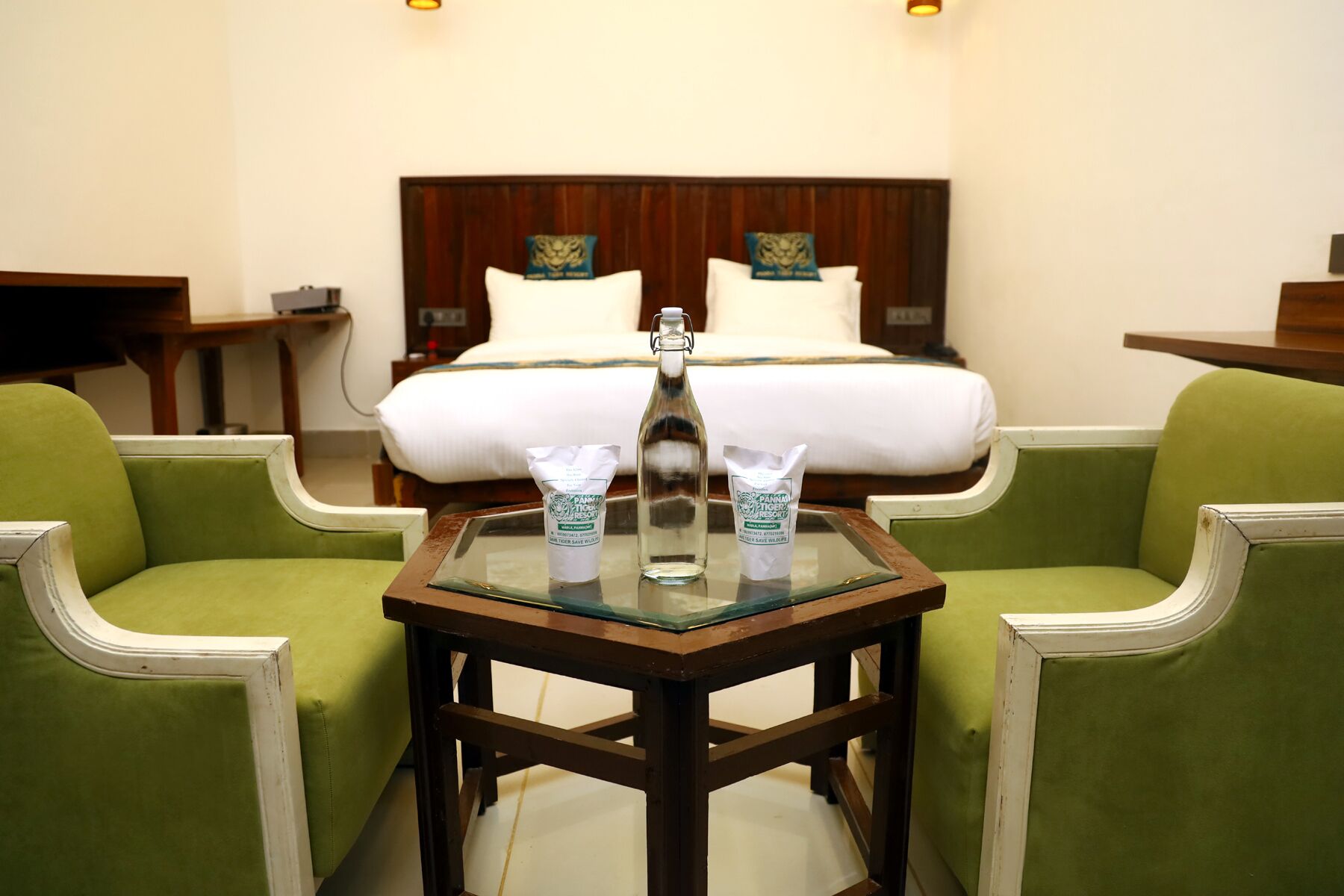Habitación de hotel con cama, dos sillones verdes, una mesita con una botella y dos paquetes, y cabecero de madera adornado con almohadones.