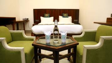 Habitación de hotel con cama, dos sillones verdes, una mesita con una botella y dos paquetes, y cabecero de madera adornado con almohadones.