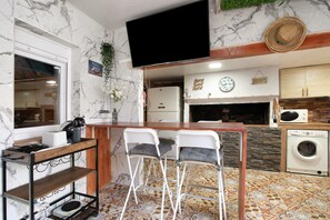 A compact kitchen with a marble backsplash, a flat-screen TV, a bar area with stools, a washing machine, and a clock.