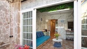 A stone exterior wall, a glass door leading to a room with a blue sofa, a tiled floor, and a small round table with a vase. - Holiday Home "Casa Tenerife Primera Linea" with Sea Views, 3 Private Terraces and Wi-Fi (tenerife)
