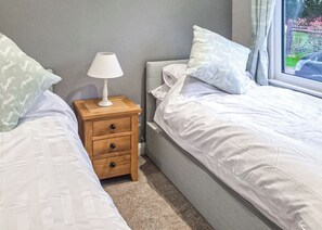 A bedroom with two beds, a bedside table with a lamp, and a window with a view of greenery. - Rabbit Lodge is designed to provide you with a luxurious and comfortable stay. (Thorney, Nr Lincoln)