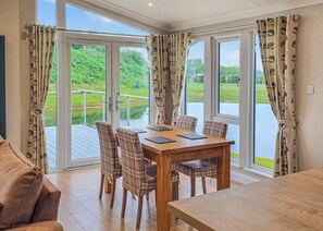 A dining area with a table and chairs, large windows with curtains, and a view of greenery and water. - Rabbit Lodge is designed to provide you with a luxurious and comfortable stay. (Thorney, Nr Lincoln)