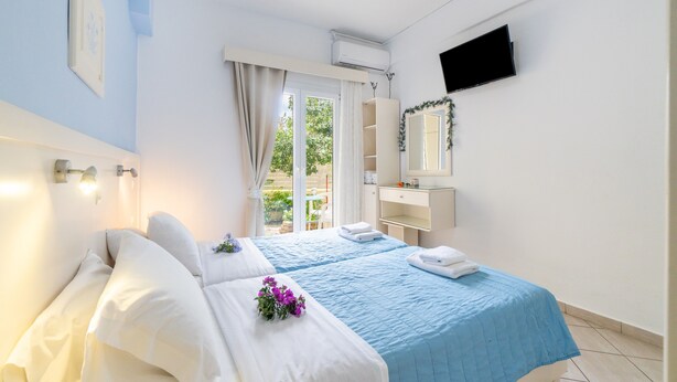A hotel room with two beds, a TV, a window with curtains, a bathroom, and a view of greenery outside.