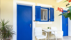 A room with a blue door, a white table with two chairs, a small potted plant, and a window with blue shutters.