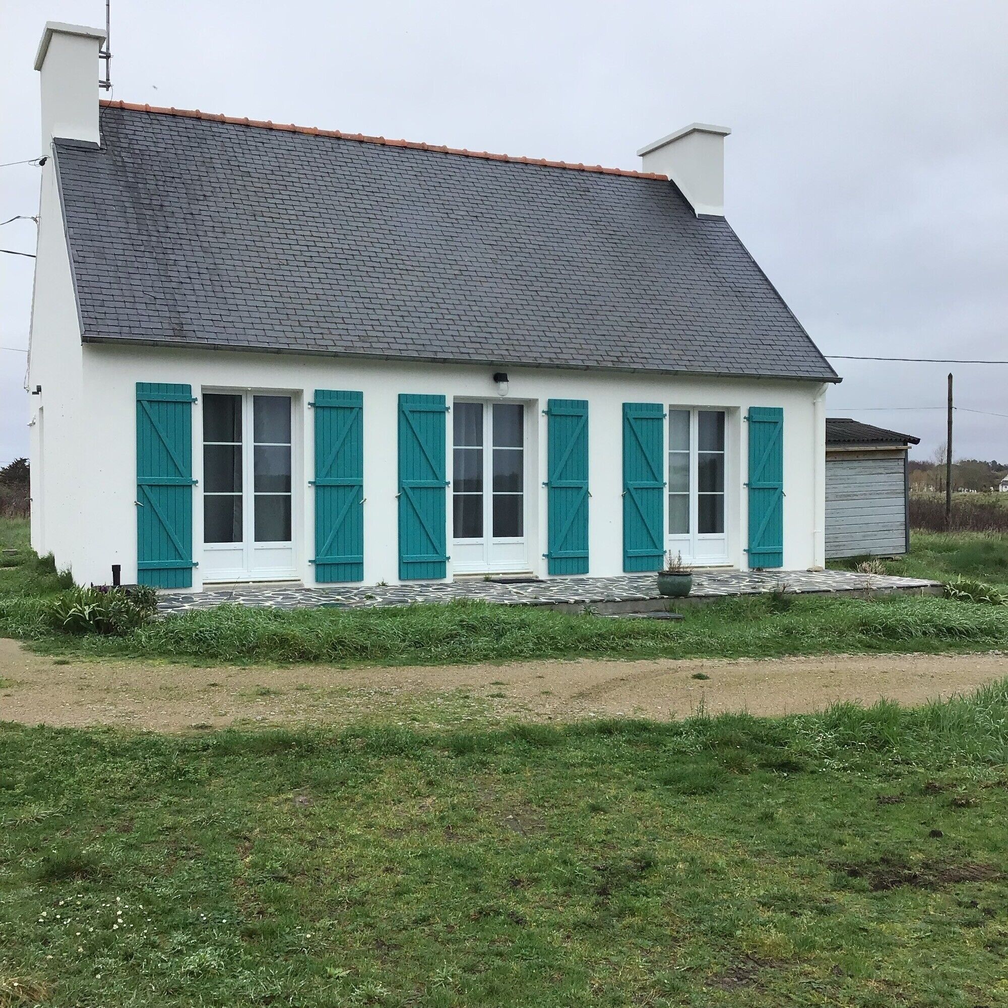 A white building with teal shutters, a grey roof, and a stone foundation.