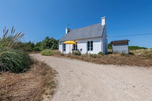 Une maison blanche avec une allée en gravier, un parasol jaune et un petit abri de jardin.