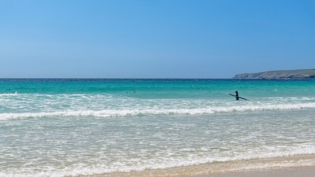 Una playa con agua cristalina, una persona practicando surf y acantilados a lo lejos.