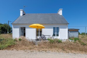 Une maison blanche avec un toit gris, un parapluie jaune, et une petite table et une chaise à l’extérieur.