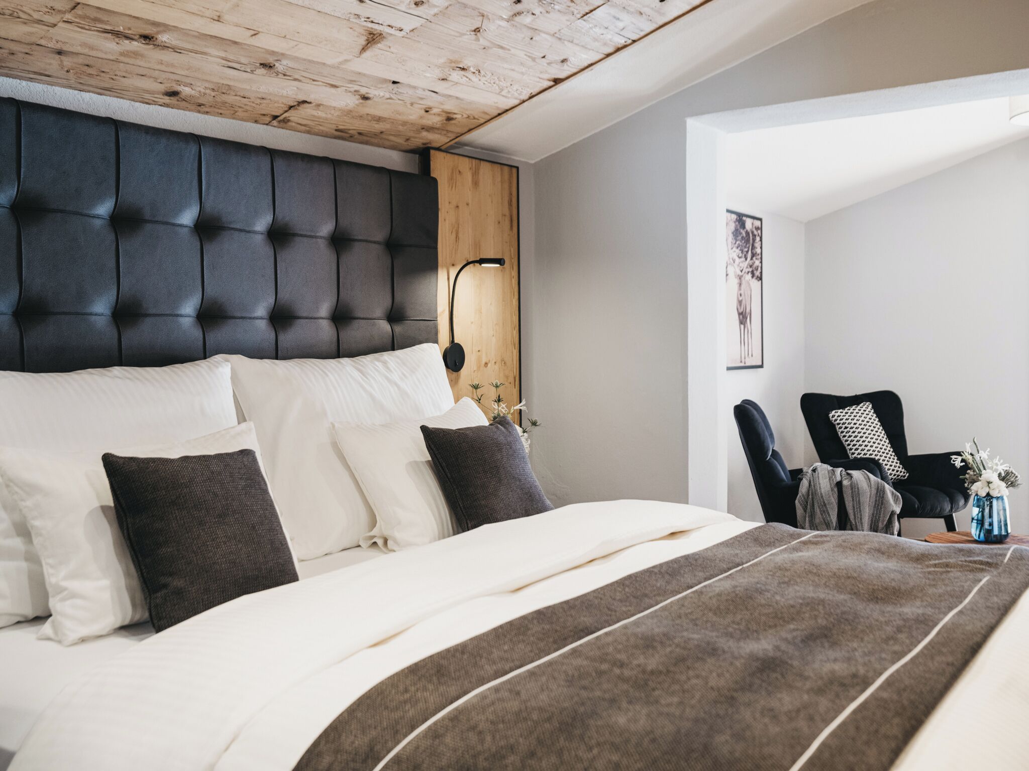 A bedroom with a tufted headboard, a bed with white and gray bedding, a wooden ceiling, a black chair with a cushion, and a wall-mounted lamp.