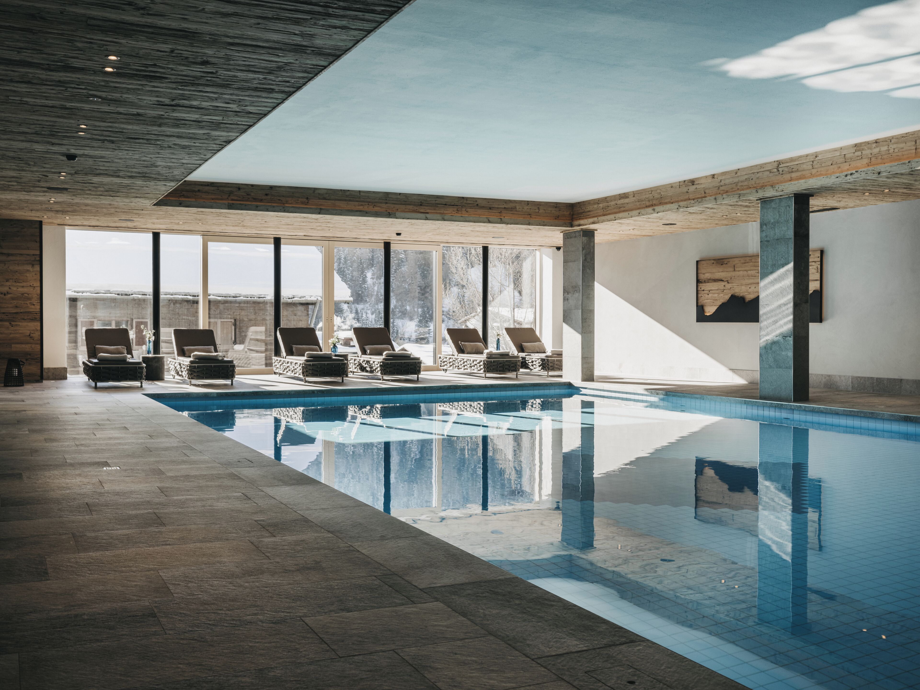 A modern indoor swimming pool with clear glass walls, wooden beams, and lounge chairs.
