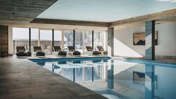 A modern indoor swimming pool with clear glass walls, wooden beams, and lounge chairs.