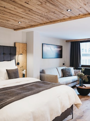 A modern hotel room with a wooden ceiling, a large bed, a sofa, a side table with a vase, and a painting on the wall. - VAYA Nauders (Nauders)