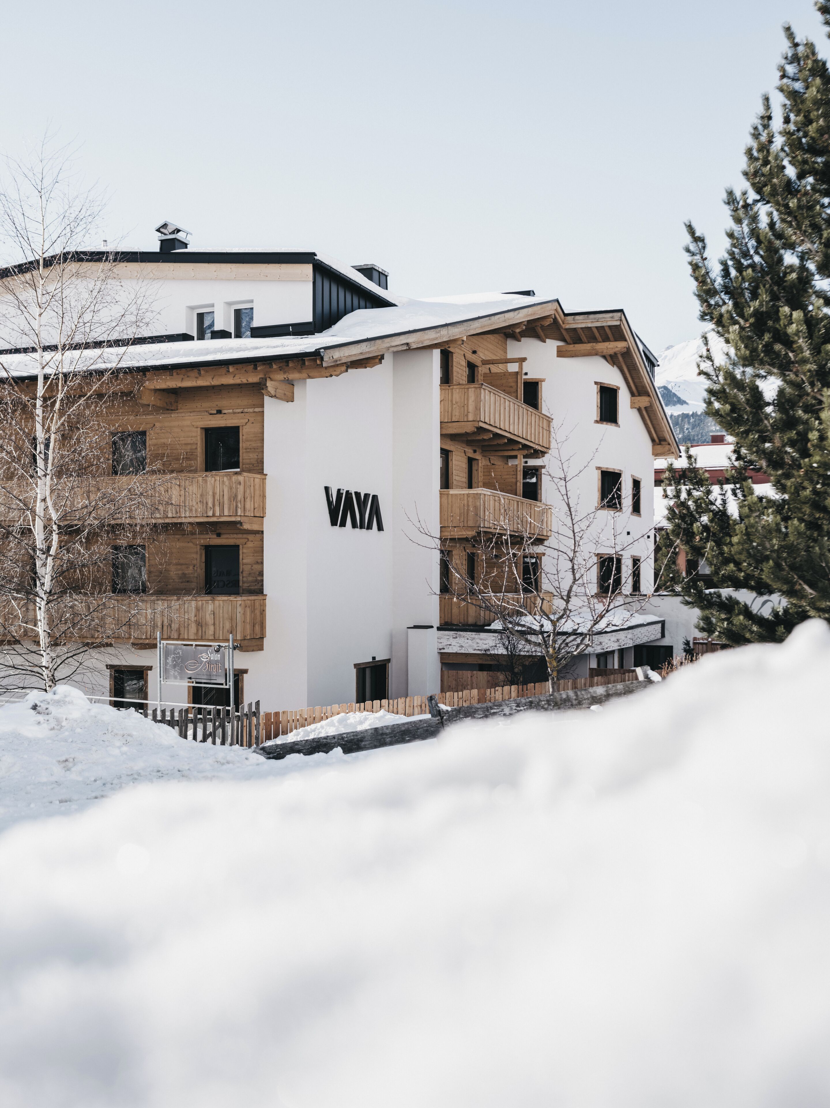 A modern multi-story building with the name "VAYA" prominently displayed. It features wooden balconies and a snow-covered landscape.