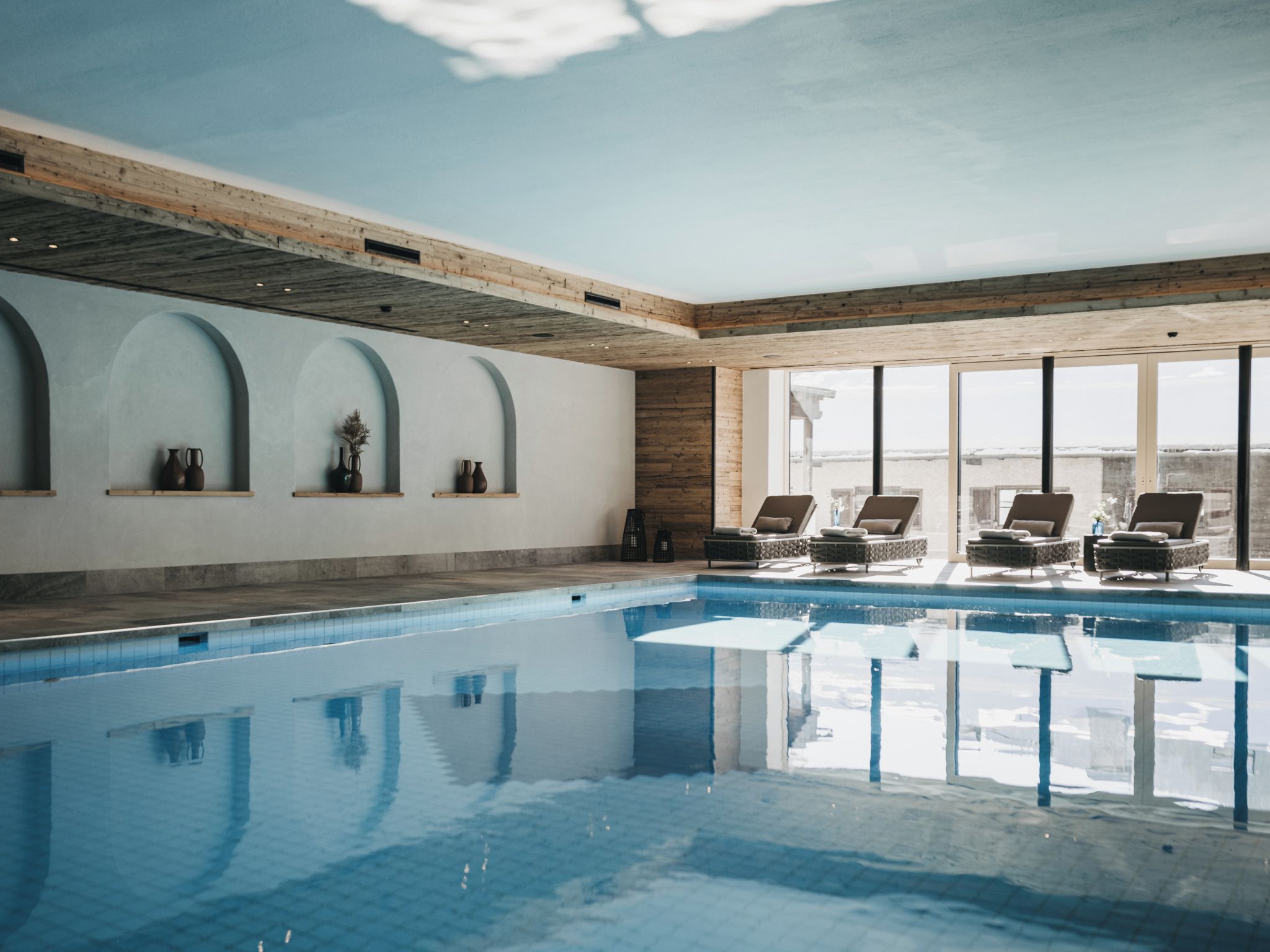 An indoor swimming pool with clear blue water, wooden beams on the ceiling, and lounge chairs by the poolside.
