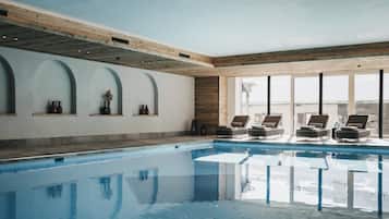 An indoor swimming pool with clear blue water, wooden beams on the ceiling, and lounge chairs by the poolside.