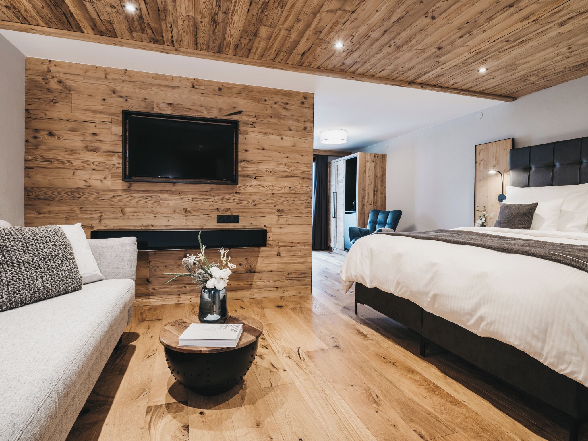 A modern bedroom with a wooden ceiling, a flat-screen TV mounted on the wall, a bed with white bedding, a dark headboard, and a small round table with a vase of flowers.