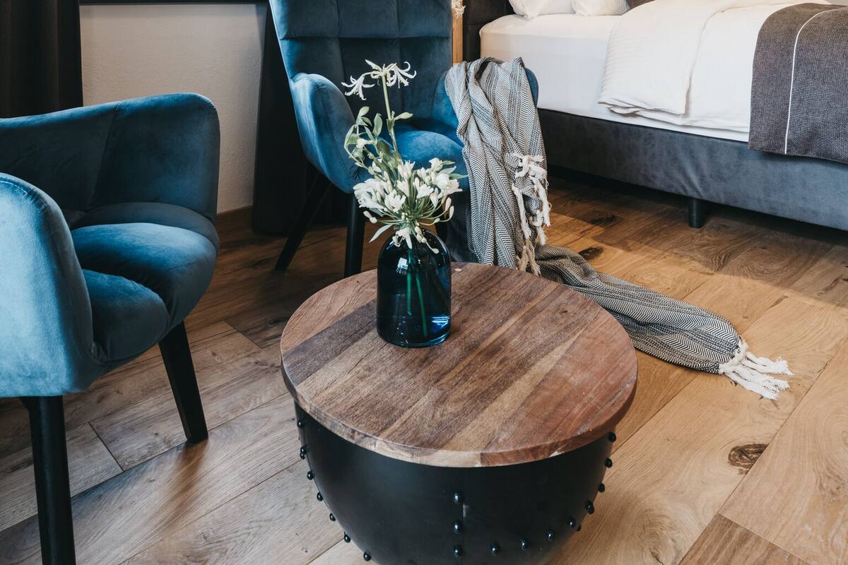 Un dormitorio moderno con techo de madera, una cama grande, un sillón azul, una mesa de centro redonda con un florero y vistas a edificios a través de una ventana.