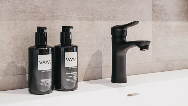 A bathroom with a modern design, featuring a grey tiled wall, a black faucet, and two VAYA hand and body lotion bottles. - VAYA Nauders (Nauders)
