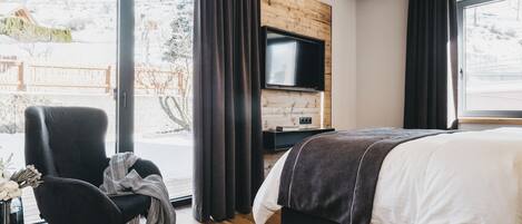 A modern hotel room with a large bed, a TV mounted on a wooden wall, and a view of a snowy landscape through large windows.