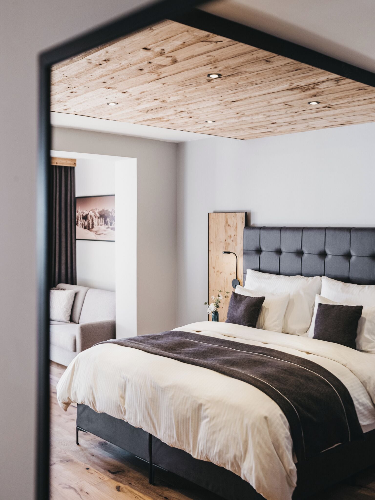 Une chambre à coucher moderne avec un plafond en bois, un grand lit doté d’une tête de lit rembourrée de couleur grise et une table de chevet sur laquelle est posé un vase.