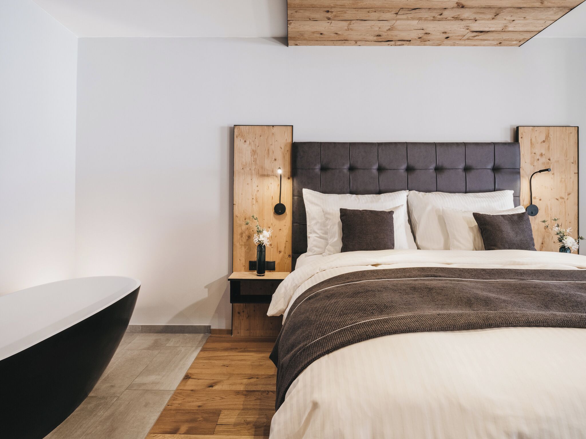 Une chambre moderne avec une tête de lit en bois, un lit avec une literie blanche et foncée, une table de chevet ornée d’un vase et un plancher en bois.