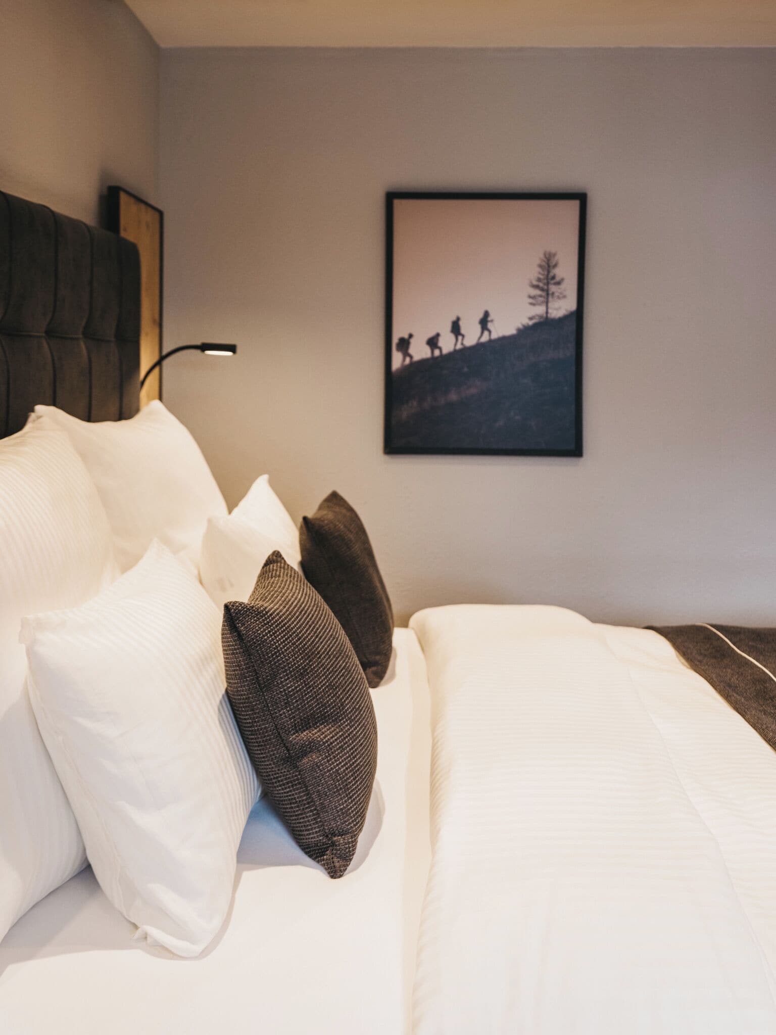 A neatly made bed with white and brown pillows, a headboard, and a wall-mounted picture of hikers on a mountain.